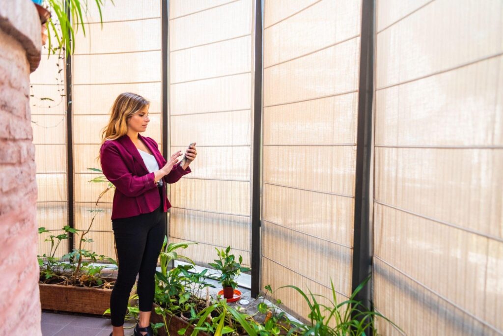 Woman adjusting smart outdoor shade settings on her phone near fabric roller shades in a sunlit indoor-outdoor space—highlighting smart shade technology and materials.