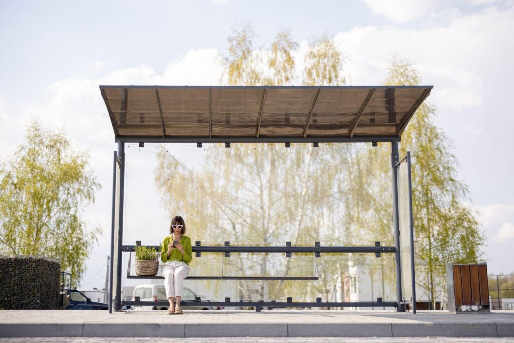 Woman sitting under a sleek metal and glass exterior shade structure at a bus stop, showing functional and energy-saving outdoor shade solutions